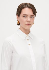 Hvit skjorte med peke-krage, som har tre gull dekorative knapper og en jevn, ensfarget tekstur. Gull øredobber.