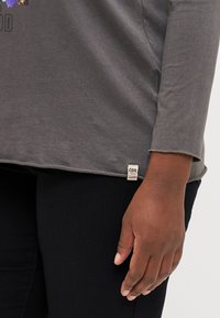 Grå långärmad tröja med rundad nederkant, med en liten etikett i botten. Slät textur med subtila sömnadsdetaljer.