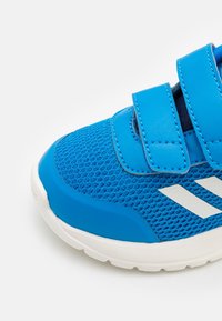 Chaussure de sport bleue avec une tige en maille, des bandes en cuir synthétique lisse et des rayures blanches. Semelle en caoutchouc épais pour une meilleure adhérence.