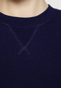 Närbild av en marinblå sweatshirt med rund hals och en broderad "X"-detalj nedanför kragen på en persons hals och övre bröst.