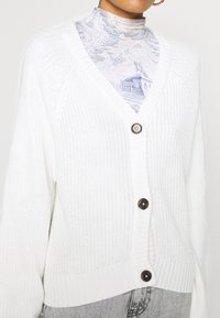 Cárdigan blanco de punto con escote en V, que cuenta con tres grandes botones marrones, dobladillo y puños acanalados, y un patrón texturizado.