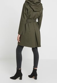 Manteau vert olive, mi-long avec capuche, taille ceinturée et détail de pli au dos, associé à des bottines noires et un jean skinny foncé.
