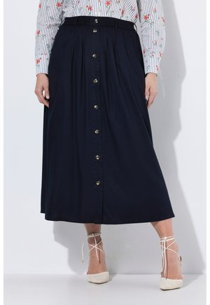 Kobieta w granatowej, zapinanej na guziki spódnicy midi, w paskowaną kwiecistą bluzkę oraz białe sznurowane szpilki z ostrym noskiem.