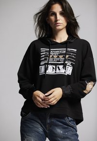 Black hooded sweatshirt with graphic print featuring white text and design details. Cuffed sleeves and relaxed fit. Worn with distressed denim.