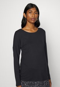Blusa de manga longa preta em tecido suave, com um decote largo e um corte descontraído. O modelo combina-a com uma saia estampada.