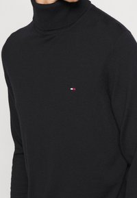 Pull noir à col roulé avec un petit logo rectangulaire rouge, blanc et bleu sur le côté gauche de la poitrine, porté par une personne.