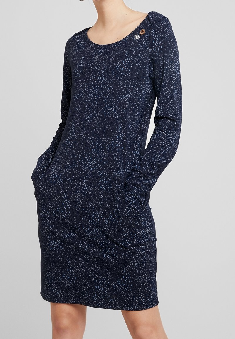 Femme portant une robe bleu marine à manches longues avec de petites taches bleu clair et des poches, debout devant un fond gris clair uni.