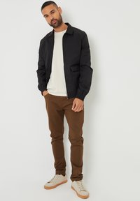 Giacca bomber nera con polsini a coste, t-shirt beige, pantaloni marroni e sneakers bianche con suole color marrone chiaro, che mostrano un design di abbigliamento casual.