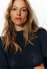 Pull en tricot marine avec un motif en dentelle et un col haut. Le modèle a de longs cheveux blonds ondulés et un look de maquillage naturel. Contexte simple.