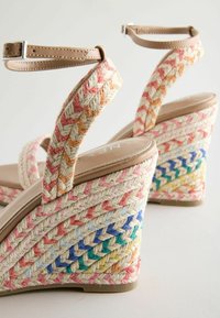 Sandales compensées avec des détails en corde tressée multicolore, des brides de cheville beiges avec boucles et des semelles couleur tan sur fond blanc.