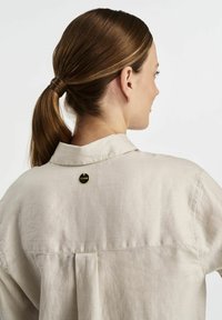 Beżowa lniana koszula z wyraźnym kołnierzem, charakteryzująca się delikatną teksturą i okrągłym, złotym guzikiem z tyłu.