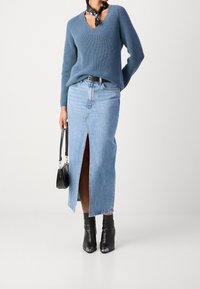 Pull en tricot bleu avec col en V, jupe en denim bleu clair avec fente devant, bottines noires et petit sac à main noir. Écharpe noire en accent.