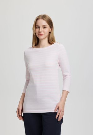 Joven mujer con cabello rubio lacio, viste una camisa de manga larga de rayas rosa claro y pantalones oscuros, de pie frente a un fondo gris liso.