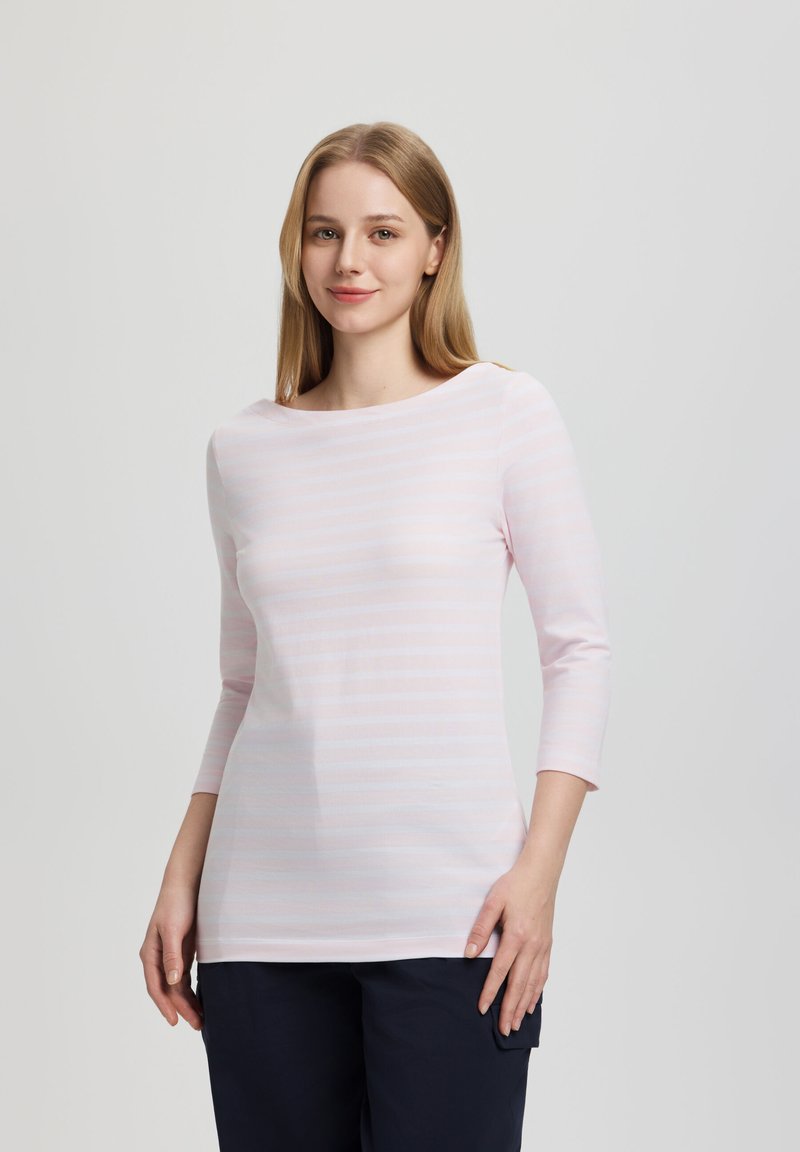 Junge Frau mit glattem blondem Haar trägt ein hellrosa gestreiftes Langarmshirt und dunkle Hosen, steht vor einem schlichten grauen Hintergrund.