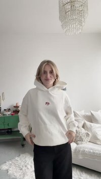 White hoodie with a front pocket and red embroidered logo. Model stands against a neutral background, wearing black pants.