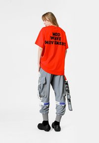 Camiseta de algodón roja con el texto negro "MOVIMIENTO MOD WAVE" en la espalda, combinada con pantalones cargo grises que presentan múltiples bolsillos y detalles.