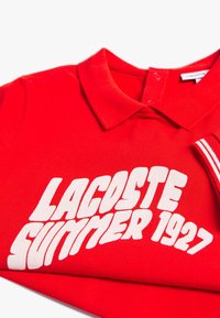 Czerwona koszulka polo z kołnierzykiem, z odważnym białym napisem "LACOSTE SUMMER 1927" na przodzie oraz paskowanym detalem na rękawie.