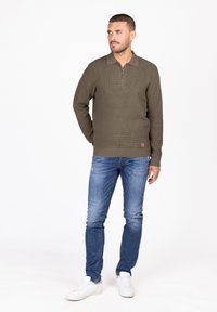Olivgrüner Reißverschluss-Pullover aus strukturierter Stoff, kombiniert mit hellblauen, verwaschenen Jeans und weißen Sneakern. Einfaches, lässiges Design.