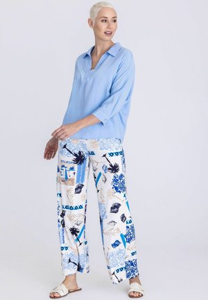 Frau mit kurzen blonden Haaren, die eine hellblaue Bluse, weiße Hosen mit weitem Bein und blauen Palmen- und Muschelmustern sowie weiße Sandalen trägt.