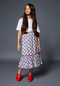 Camicia bianca a maniche corte, gonna a fantasia floreale con fiori rossi e blu, orlo a balze a strati e zoccoli rossi vivaci. Sfondo grigio.