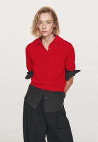 Una donna con i capelli biondi corti indossa un maglione polo rosso a maniche corte sopra una maglietta nera a maniche lunghe e pantaloni neri larghi, in piedi davanti a uno sfondo bianco.
