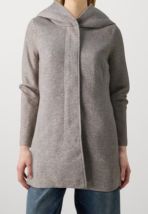 Frau trägt einen grauen Kapuzenmantel mit langen Ärmeln und verdecktem vorderem Verschluss, kombiniert mit blauen Jeans, vor einem einfarbigen hellen Hintergrund.