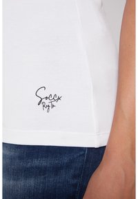Camiseta blanca de algodón con dobladillo redondeado y un logo negro de "Soccx" cerca de la parte inferior derecha. Se muestra una combinación con jeans oscuros.