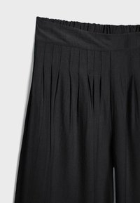 Jupe noire taille haute avec de fines plis verticaux et une large ceinture lisse sur un fond clair.