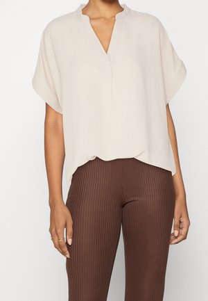 Vrouw draagt een losse beige top met korte mouwen en een V-hals, gecombineerd met aansluitende bruine ribgebreide broek met hoge taille, staand tegen een witte achtergrond.