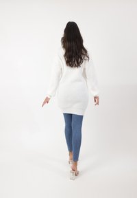 Witte gebreide oversized trui met lange mouwen, gecombineerd met strakke blauwe jeans en sandalen met doorzichtige hakken, vanuit de achterkant bekeken.