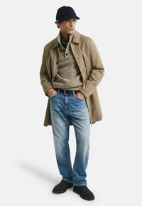 Mann in beiger Jacke, braunem Pullover, blauen Jeans, schwarzen Stiefeln und einer marineblauen Mütze, der mit den Händen in den Taschen steht.