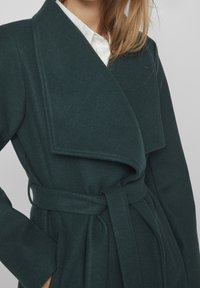 Tumma vihreä villasekoitekääreetakki, jossa on leveä kaulus, vyötärölle solmittava vyö ja sivutaskut. Kankaan pinta on pehmeä ja teksturoitu.