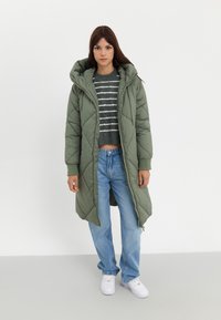Manteau matelassé long, de couleur vert olive, avec une fermeture éclair et une capuche réglable, porté sur un pull court rayé et un jean bleu clair.