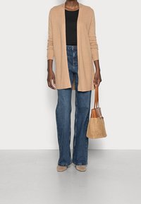 Long gilet beige sur un haut noir, associé à un jean évasé bleu. Accessoirisé avec un sac tressé marron clair et des chaussures beiges.