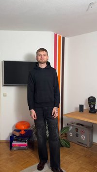 Μαύρο φούτερ με κουκούλα, μαύρο τζιν με κεντήματα, καφέ παπούτσια. Απλός σχεδιασμός, άνετη εφαρμογή. Πολύχρωμος ριγέ τοίχος στο φόντο με διακοσμητικά αντικείμενα.