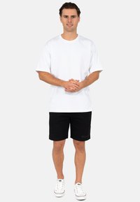 Witte T-shirt met korte mouwen, zwarte shorts en witte sneakers. T-shirt heeft een ronde halslijn, de shorts zijn textuur en bevatten een klein logo.