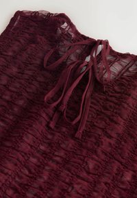 Burgundy lace top featuring a textured pattern and a tie closure at the neck. The fabric is lightweight and semi-sheer.