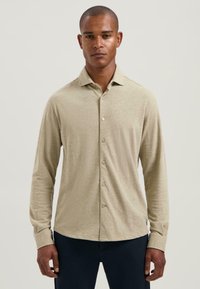Man met een beige langemouwen overhemd met knopen en donkere broek, staande tegen een effen lichte achtergrond, recht vooruit kijkend.