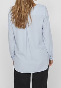 Blouse bleu clair à manches longues avec poignets boutonnés, ourlet arrondi et détail d'empiècement au dos. Le tissu semble soyeux et lisse.