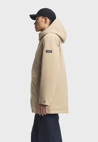Beige waterafstotende jas met een capuchon, drukknoopsluiting en een zwart logo patch op de linker mouw. Draagt het in combinatie met donkere broeken en een zwarte pet.