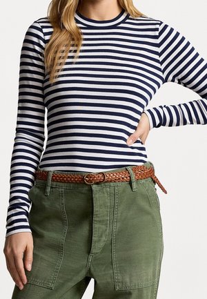 Femme portant un haut à manches longues à rayures bleu marine et blanches, un pantalon vert olive et une ceinture tressée marron, main sur la hanche devant un fond uni.
