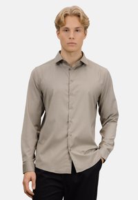 Lys brun langermet skjorte med en subtil glans, knappet foran og klassisk krage, kombinert med mørke bukser. Jevn stofftekstur.