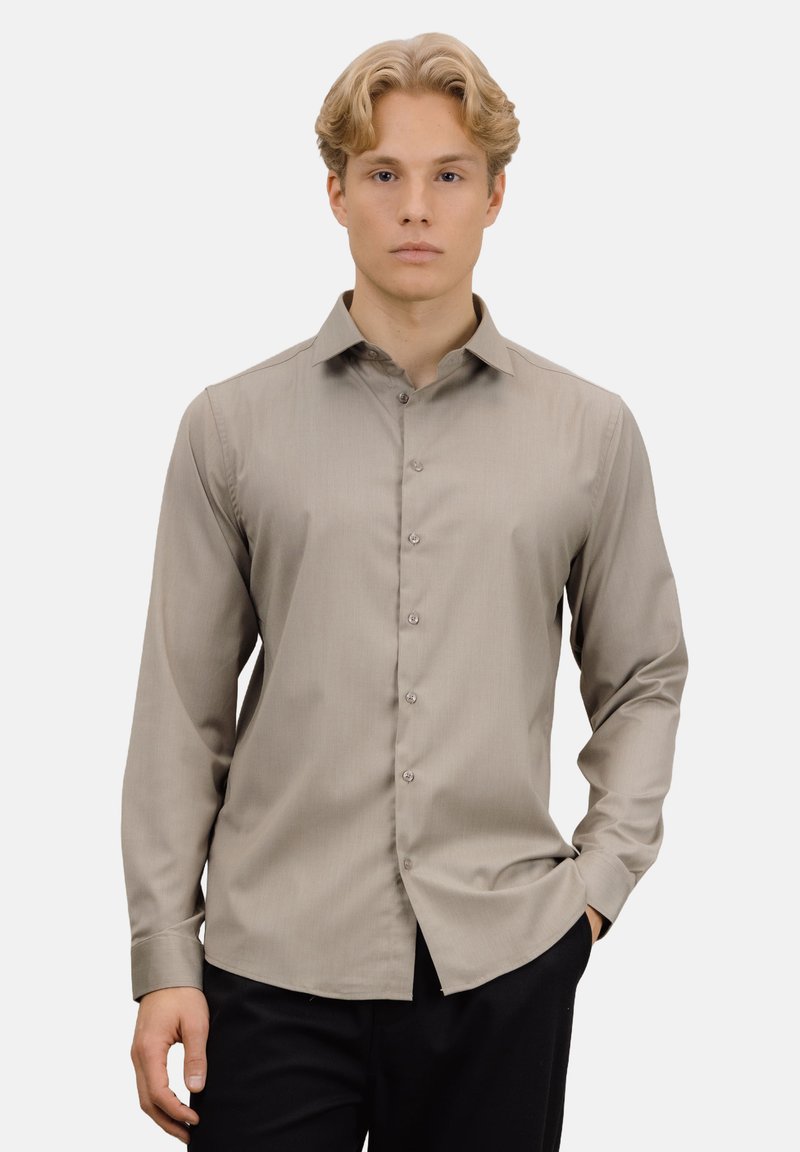 Lys brun langermet skjorte med en subtil glans, knappet foran og klassisk krage, kombinert med mørke bukser. Jevn stofftekstur.
