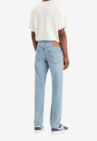Personne portant un jean droit bleu clair, un t-shirt blanc et des baskets bleues et blanches, vue de dos sur un fond blanc.