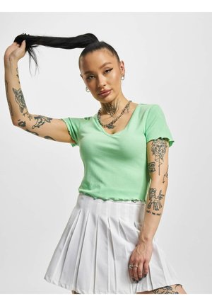 Donna tatuata con top a maniche corte verde menta e gonna bianca plissettata tiene alta la sua lunga coda di cavallo, rivolta in avanti contro uno sfondo semplice.