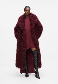 Burgundy nepbontjas met een brede kraag, lange mouwen en een textuur, gedragen over een coördinerende jurk en zwarte laarzen tot boven de dijen.