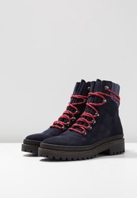 Bottes de randonnée en daim bleu marine avec des lacets rouges et blancs, une semelle en caoutchouc noire robuste, et un accent talon texturé.