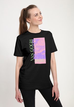 Femme aux longs cheveux attachés en queue de cheval, portant un t-shirt noir à motif Van Gogh avec des dessins violets et roses et un texte vert.