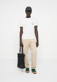 T-shirt blanc, pantalons beiges, baskets vertes avec semelles brunes et casquette noire. Tenant un sac fourre-tout noir avec des détails de logo.