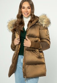 Hnědá puchová bunda s umělou kožešinou na kapuci, prošívaný design, pružné manžety a zip, nošená přes zelený svetr a modré džíny.